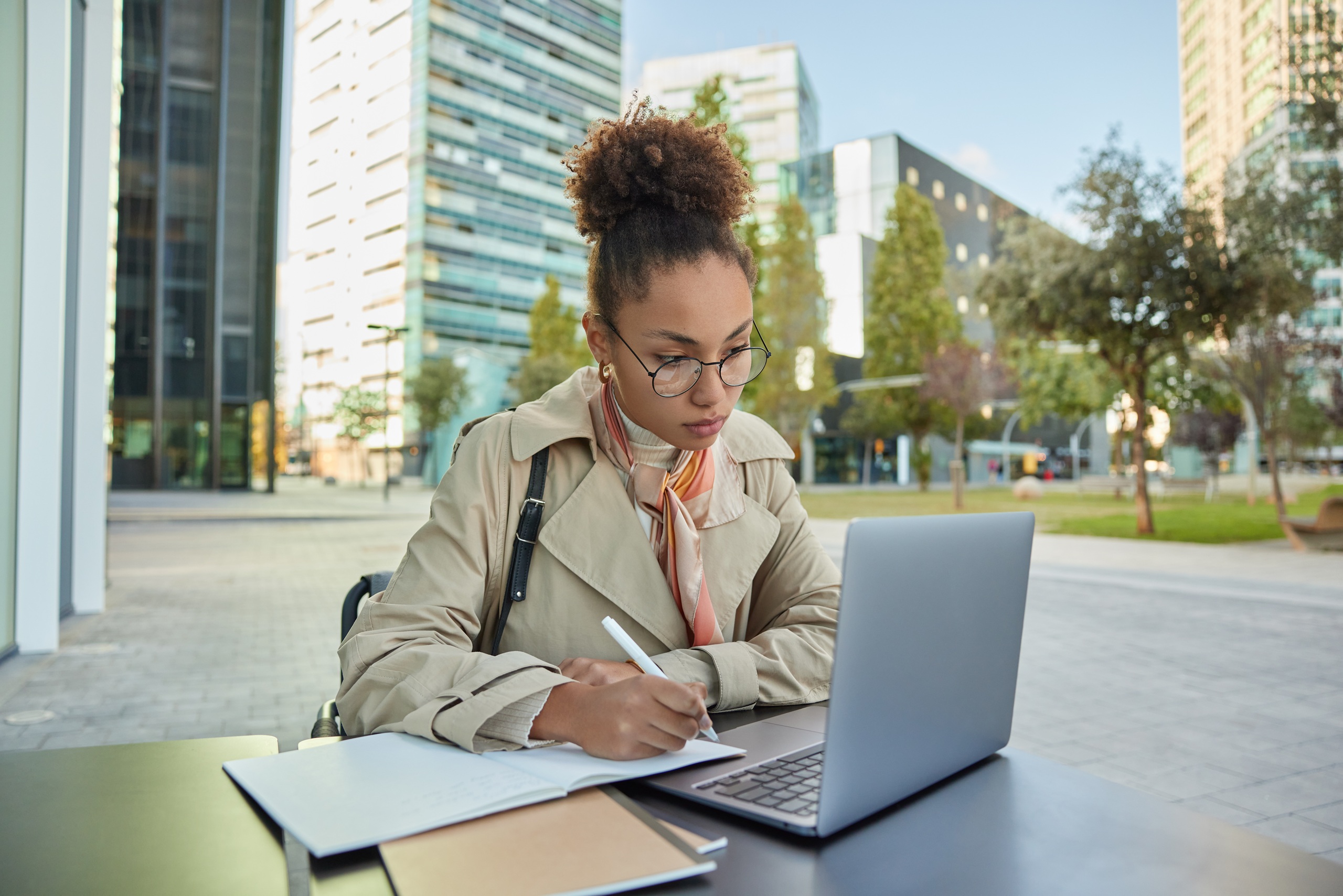 Busy student prepares for university admission watches training webinar concentrated in laptop screen wears transparent glasses and coat poses at desk outdoors. Businesswoman works online at city