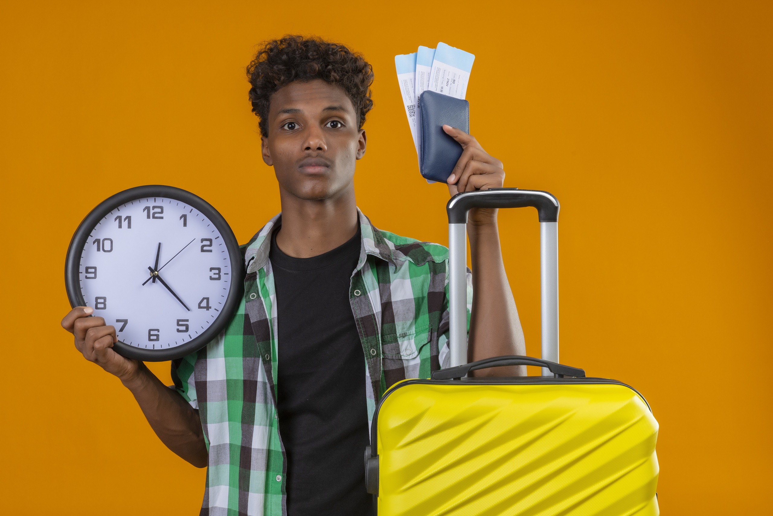 young african american traveler man standing with suitcase holding air tickets and clock looking worried and confused over orange background
